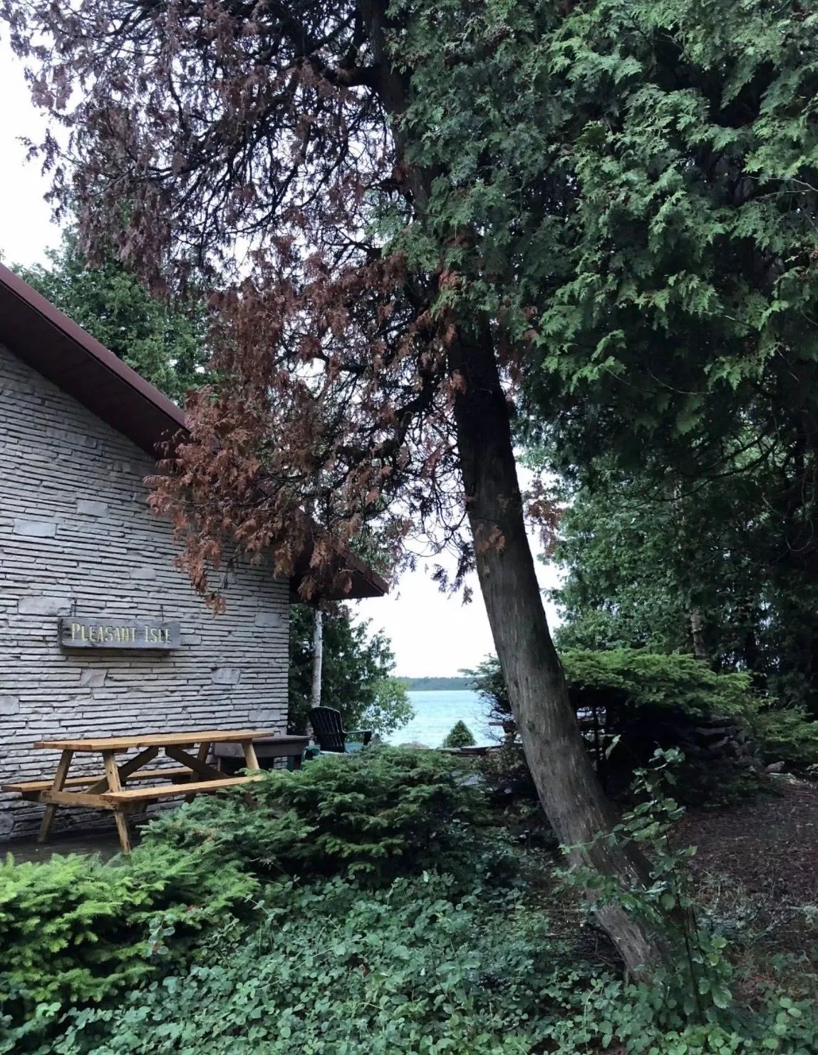 The stone cottage on the island, with a Pleasant Isle sign, a picnic table under the cedars and the lake beyond