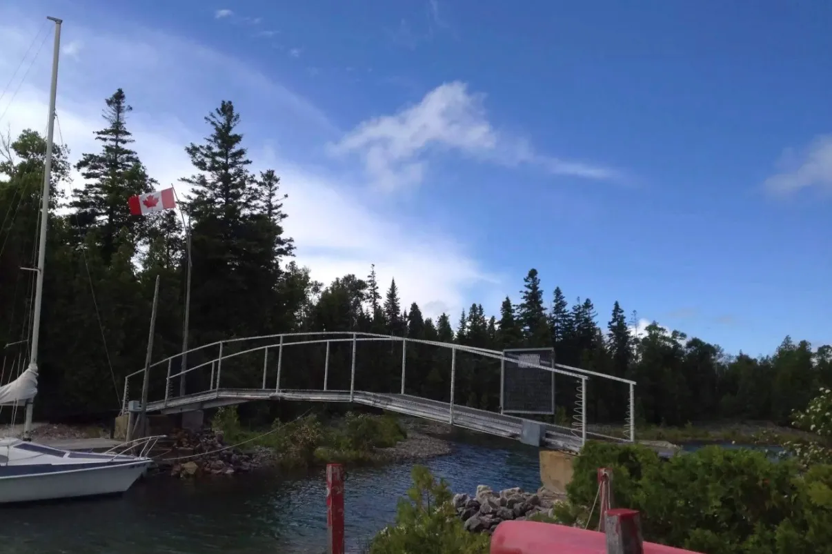 The footbridge to the island under a blue sky, with a Canadian flag and a sailboat at the dock