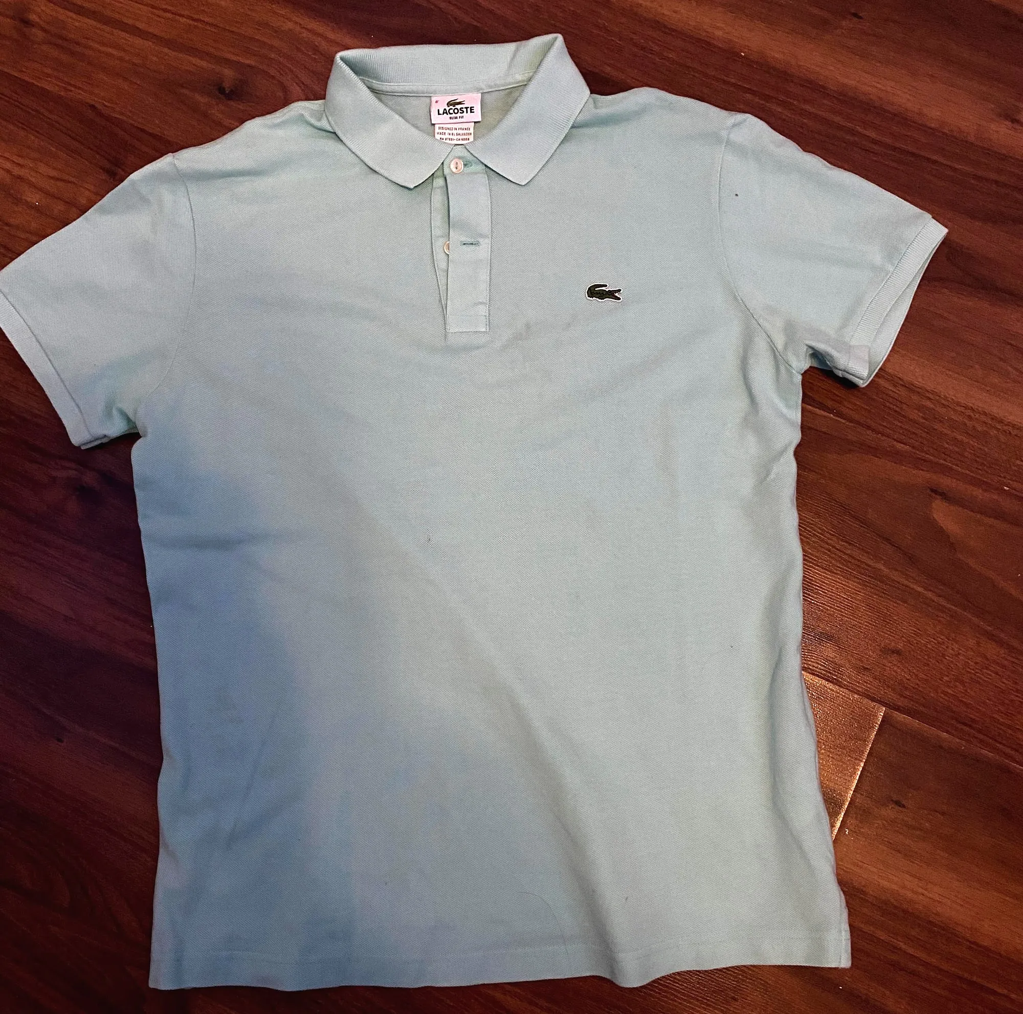 A mint green Lacoste polo shirt laid flat on a wooden floor